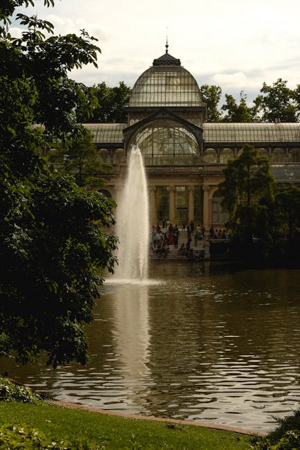 A scenic view of the Crystal Palace Park conservatory, featuring a large glass-domed structure with ornate metal framework, situated behind a pond with a prominent fountain pouring water into the lake. The surrounding area is lush with mature green trees, some branches partially framing the scene on the left. Reflection of the fountain and the conservatory can be seen in the water, which is calm aside from ripples caused by the fountain. The sky above is overcast with soft, diffused light. While the image primarily captures the park’s architecture and natural elements, it evokes the setting for home relocations or moving logistics, as seen through the peaceful environment near the conservatory, aligning with services of [COMPANY_NAME] specializing in removals within the SE19 area, such as furniture transport and packing and moving activities occurring nearby.