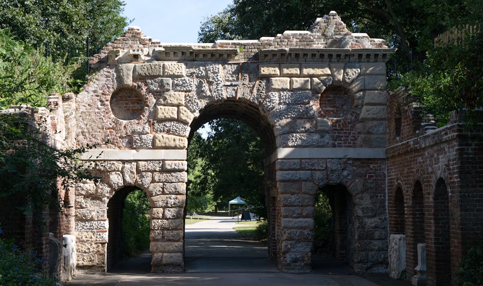 Photograph of historic brick and stone archway entrance located in Crystal Palace Park, used as part of a house removals and moving process by Removals Crystal Palace. The structure features large, weathered stone blocks with a prominent central arch and two smaller side openings, surrounded by lush green trees and foliage under a clear blue sky. In the background, a paved pathway is visible, with an area suitable for loaded vehicles and furniture transport. The archway is part of the outdoor environment, possibly serving as an access point within a park or historic site, with sunlight casting shadows on the textured brickwork. The scene illustrates the environment where home relocation activities can be coordinated, including loading furniture and packing materials, supported by equipment such as trolleys and vans, facilitating efficient and safe packing and moving operations in the SE19 area.