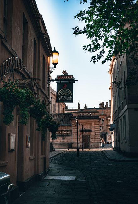 A narrow, cobbled street in Crystal Palace during daytime, flanked by historic buildings with stone facades and decorative architectural details. On the left, a building features a hanging sign for 'Fuller's', with a lamp post mounted to the wall and greenery cascading from window boxes. The street is lit by natural daylight, with shadows cast on the pavement. In the background, a larger building with classical elements such as columns and a decorative parapet is visible. The scene reflects a quiet, urban environment suitable for local house removals and furniture transport services provided by Removals Crystal Palace, especially in tight streets where careful logistics and loading processes are necessary for effective home relocation.
