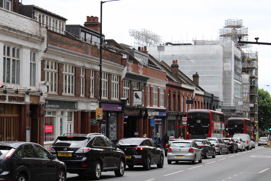 A narrow, cobbled street in Crystal Palace during daytime, flanked by historic buildings with stone facades and decorative architectural details. On the left, a building features a hanging sign for 'Fuller's', with a lamp post mounted to the wall and greenery cascading from window boxes. The street is lit by natural daylight, with shadows cast on the pavement. In the background, a larger building with classical elements such as columns and a decorative parapet is visible. The scene reflects a quiet, urban environment suitable for local house removals and furniture transport services provided by Removals Crystal Palace, especially in tight streets where careful logistics and loading processes are necessary for effective home relocation.